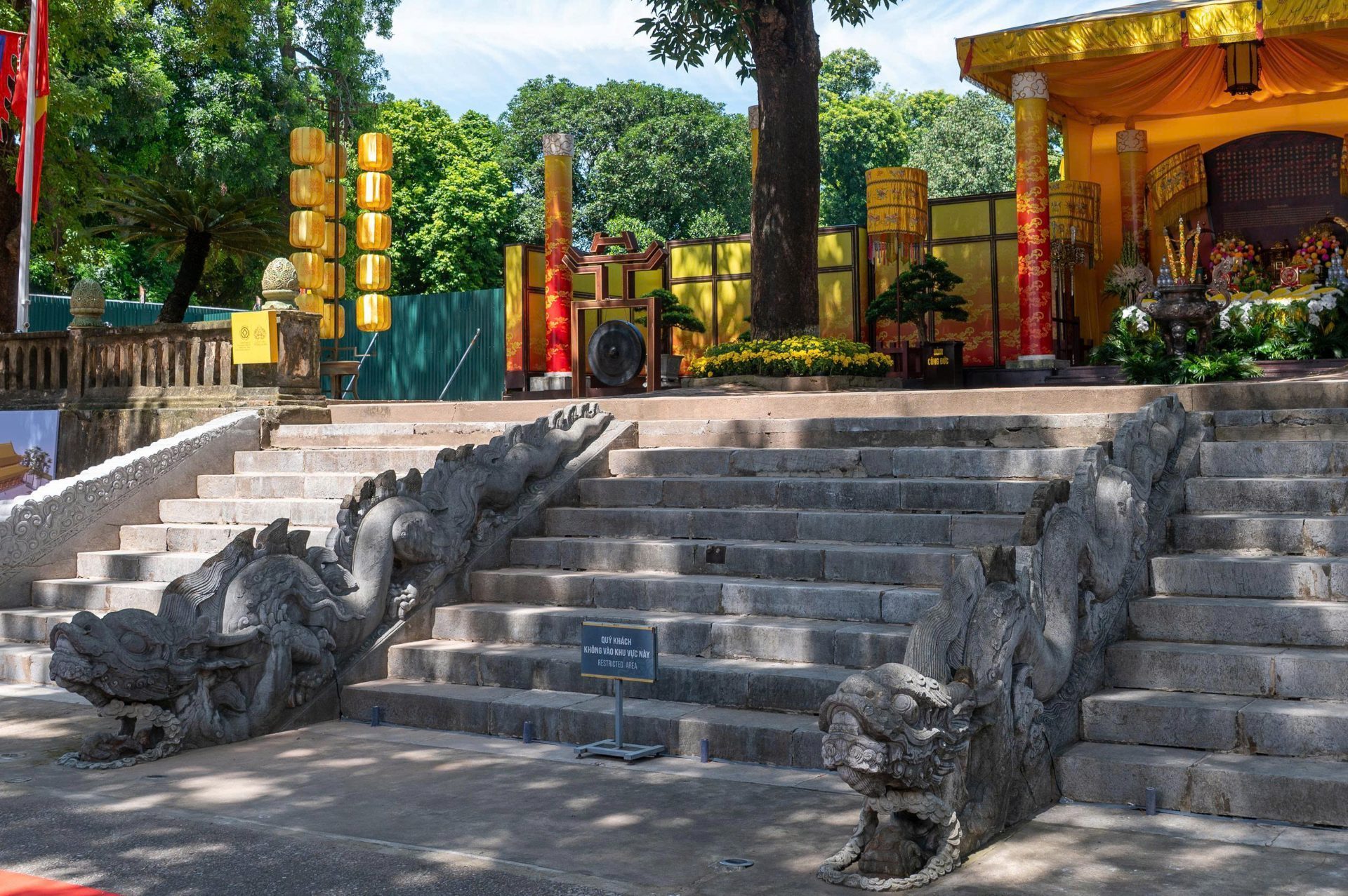 Hai con rồng đá chạm trên thành bậc, đầu ngẩng cao, thân uốn lượn dọc các bậc đá xám dẫn lên nền điện; phía sau là cây xanh và dãy nhà mái ngói.