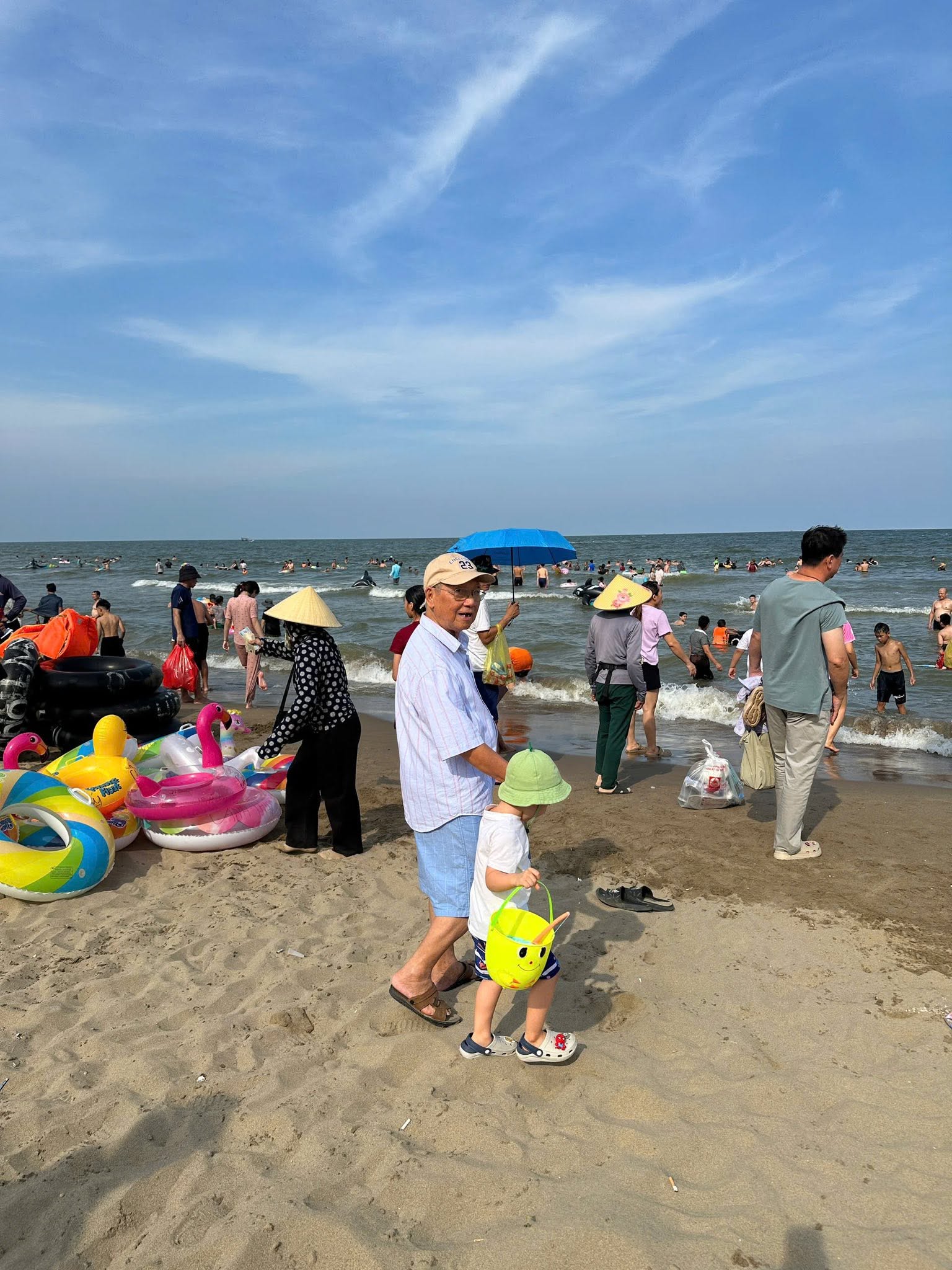 Một ông cụ dắt tay cháu nhỏ đi dạo trên bãi biển Sầm Sơn giữa những chiếc phao bơi đầy màu sắc